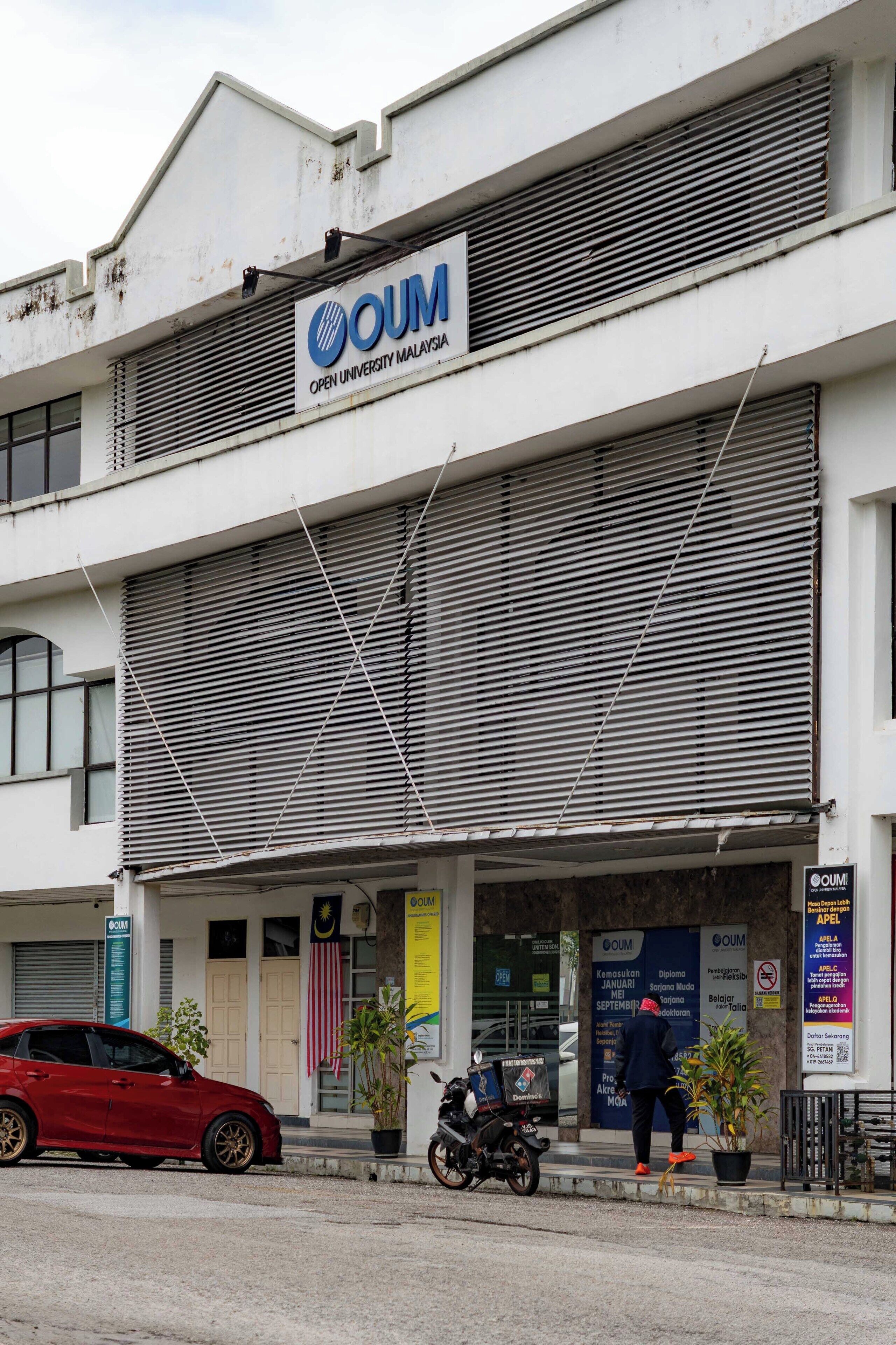 A building with a sign reading "OUM Open University Malaysia" and a red car parked in front.
