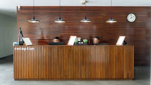 A reception desk with a wooden finish, multiple pendant lights, a clock, and a sign indicating the reception area. - T+ Hotel Sungai Petani (Sungai Petani)
