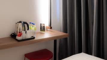 A hotel room with a wooden desk, a red stool, a white trash bin, and a bed.
