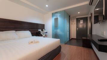 A modern hotel room with a large bed, a desk with a chair, a television, and a bathroom visible through a glass door.