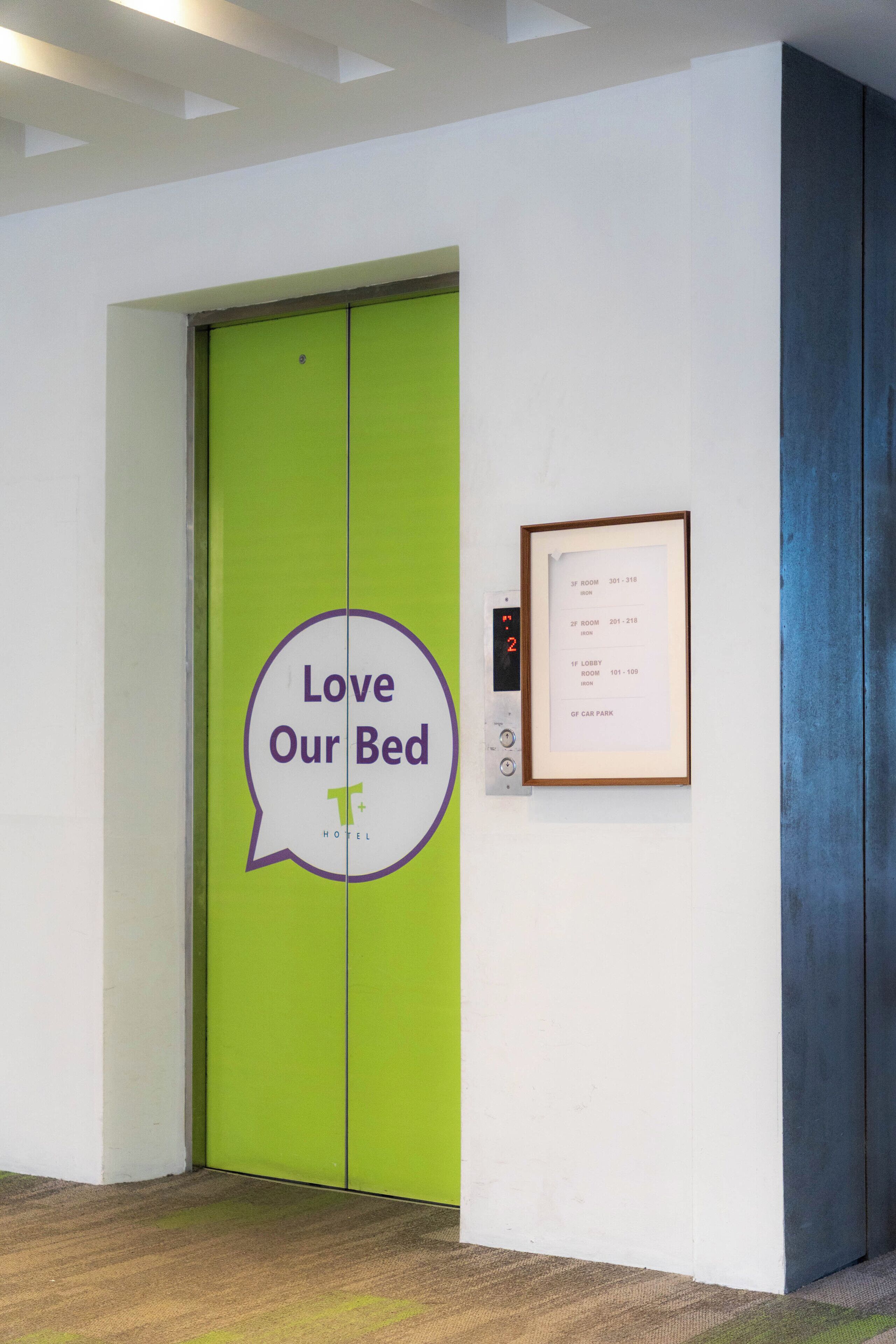 Entrada de hotel con un ascensor verde y un cartel que dice 'Love Our Bed' con un bocadillo de diálogo.