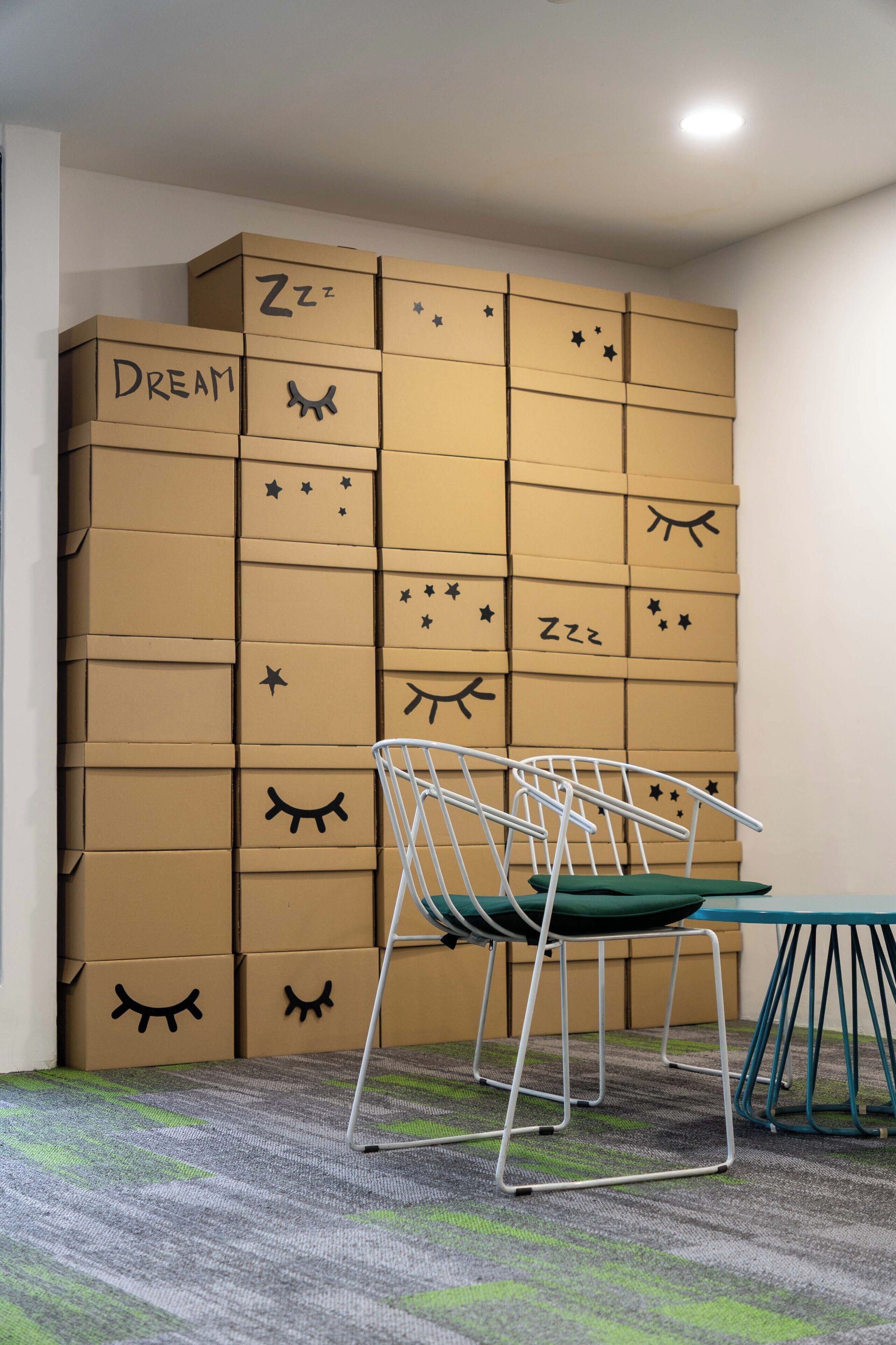 Une pièce avec une chaise en métal et une table, un sol recouvert de moquette, et un mur composé de piles de cartons ornés de divers symboles et de texte.