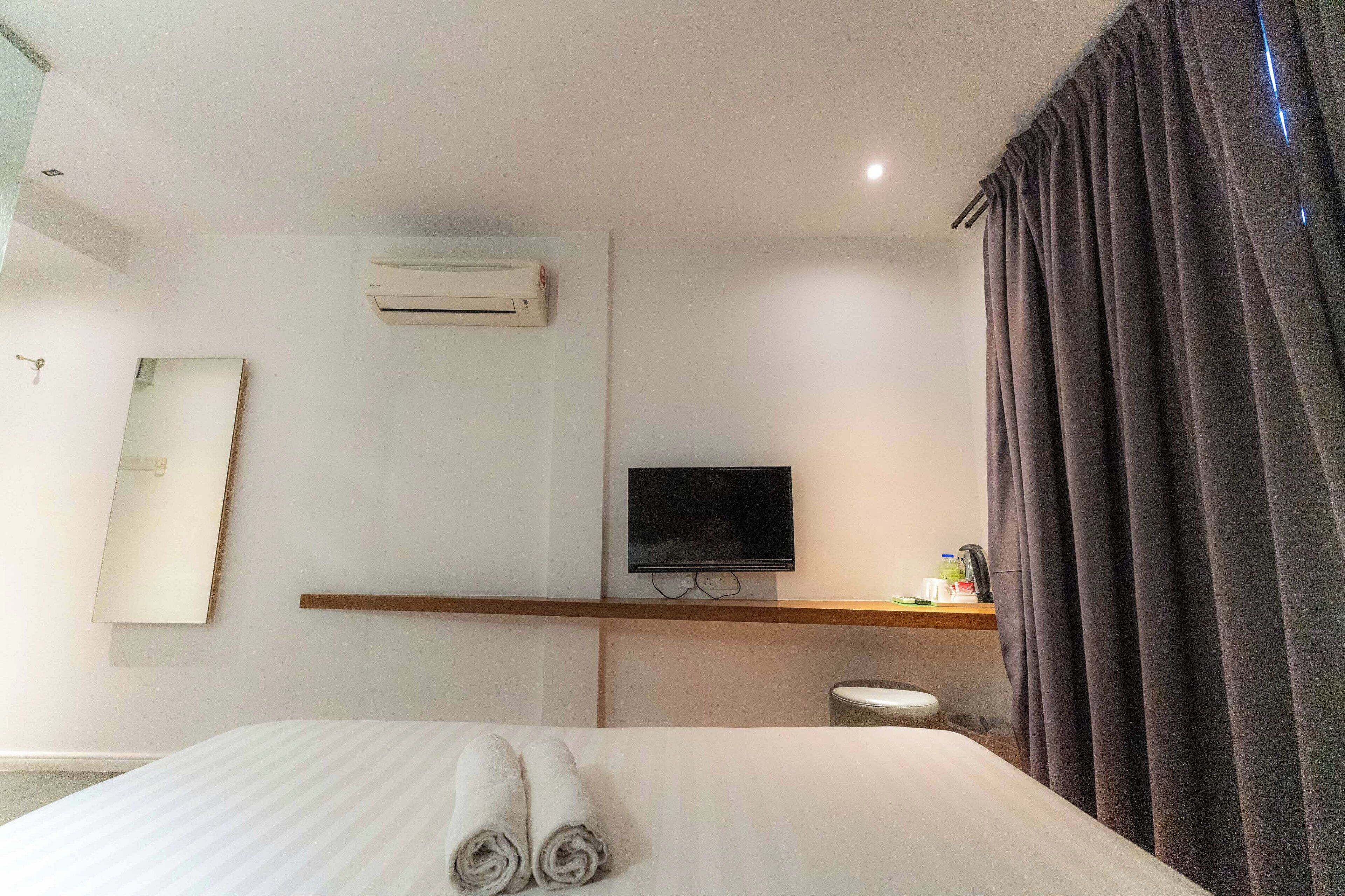 A hotel room with a bed, a flat-screen TV, an air conditioner, a small shelf with toiletries, and a curtain.