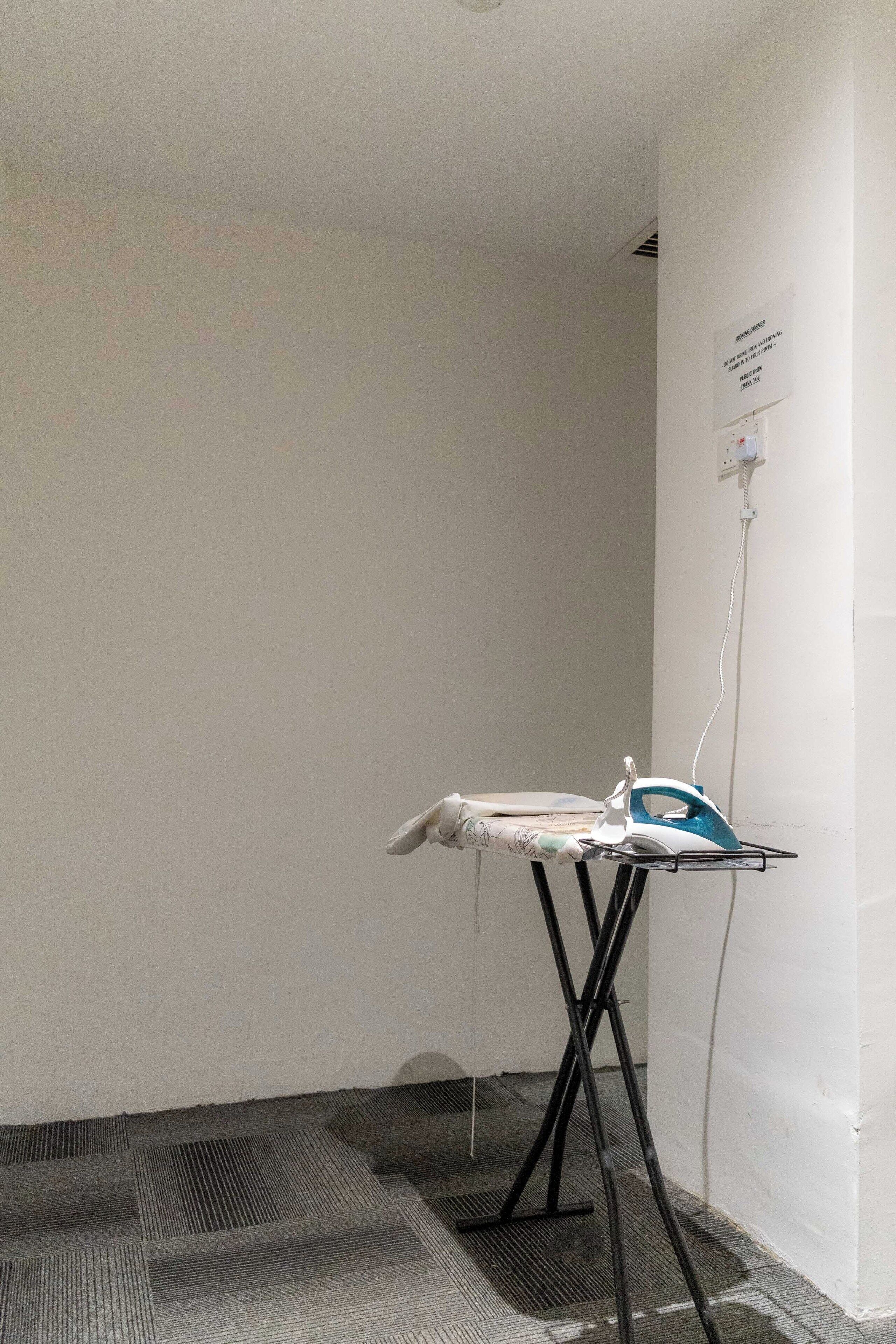 A room with a corner ironing board, a power outlet on the wall, and a patterned carpet.
