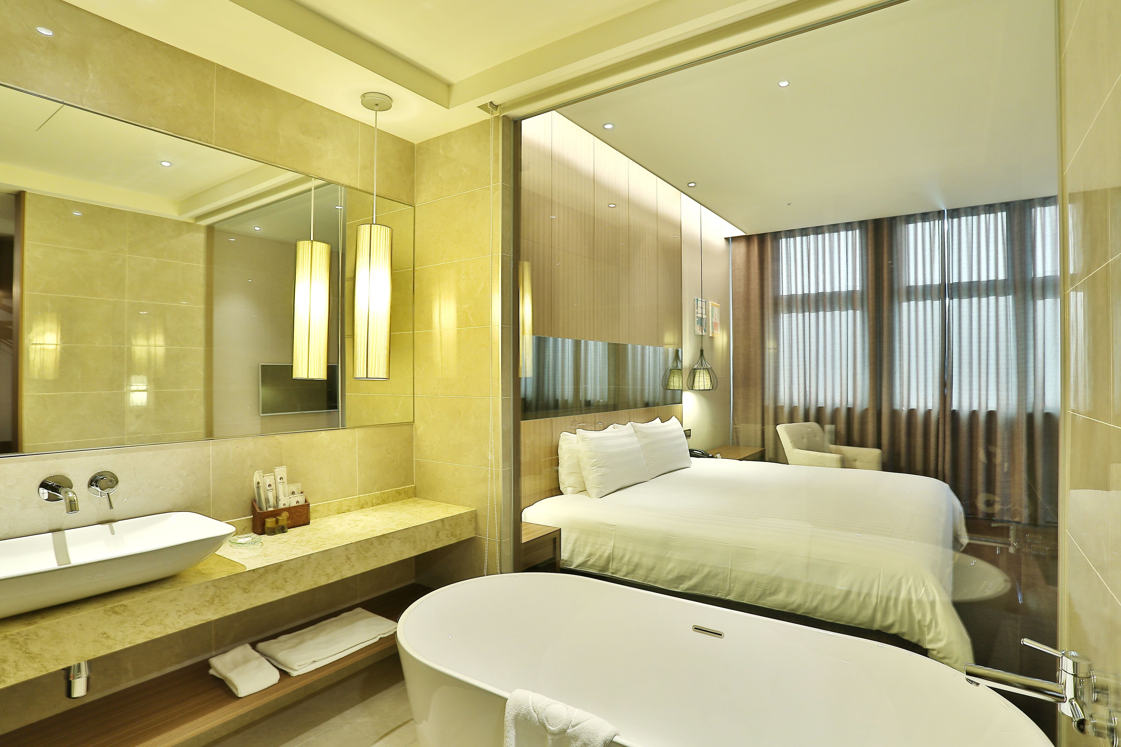 Habitación de hotel moderna con una bañera ovalada grande, una cama doble, un rincón de descanso con una silla y un espejo grande.