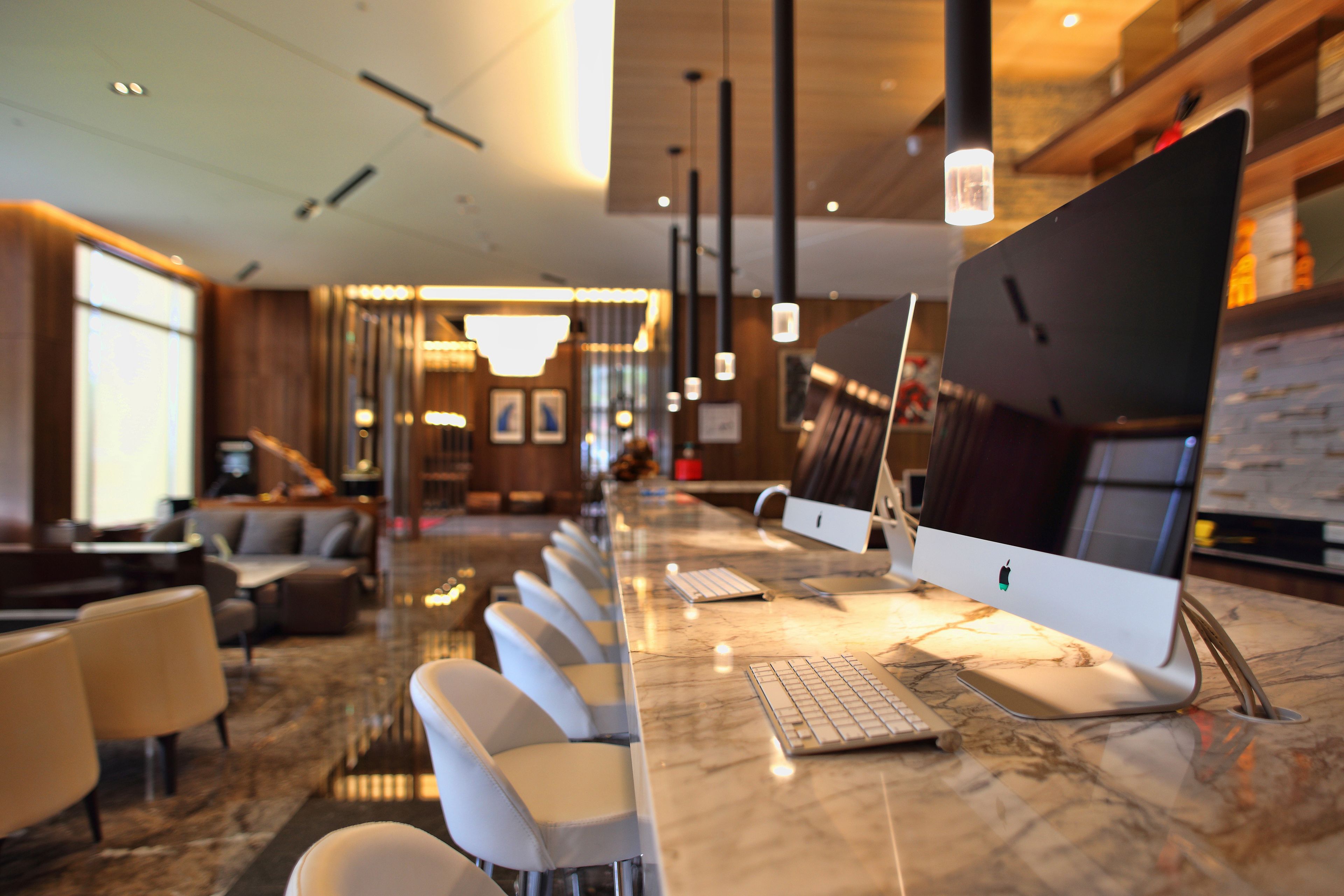 A modern hotel lobby with a long reception desk, multiple seating areas, and a large Apple iMac computer on display.