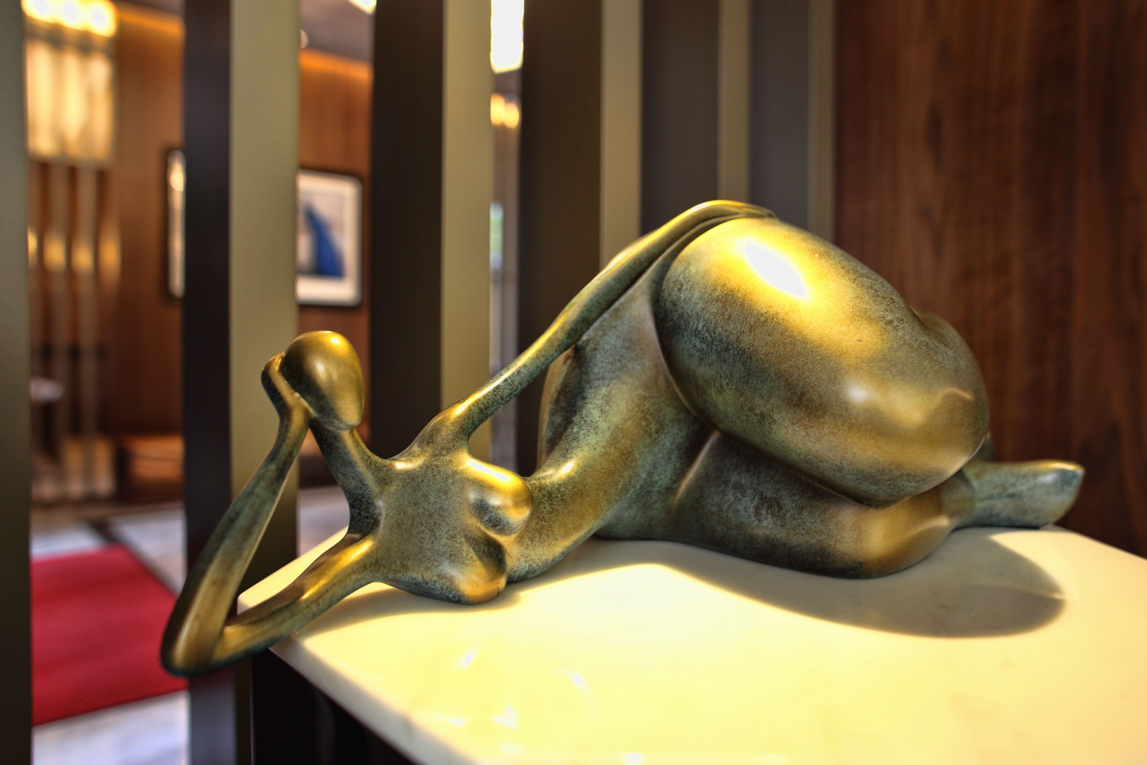 Une sculpture représentant une figure allongée sur une surface blanche, avec un arrière-plan flou comprenant un panneau en bois et un objet rouge.