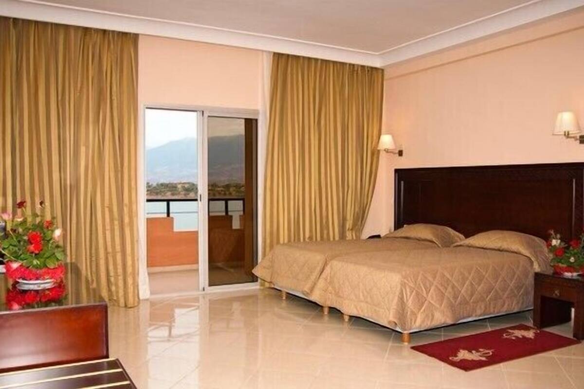 A hotel room with a bed, a desk, a chair, a nightstand, a lamp, and a view of a mountainous landscape through a sliding door.
