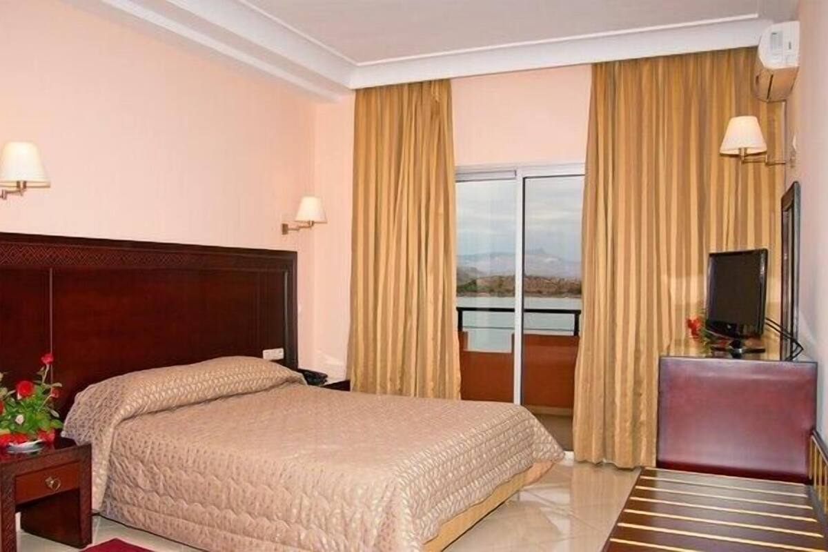 A hotel room with a large bed, a TV, a desk, and a view of the outdoors through a sliding door.