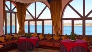 A room with a large bay window overlooking a body of water, a sofa set, and two small round tables with red tablecloths. - Hotel Chems du Lac (Bine el Ouidane)