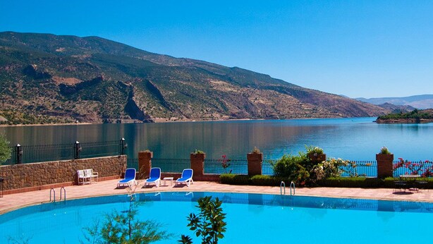 A pool with clear blue water, surrounded by a stone wall and a fence with flowers. - Hotel Chems du Lac (Bine el Ouidane)