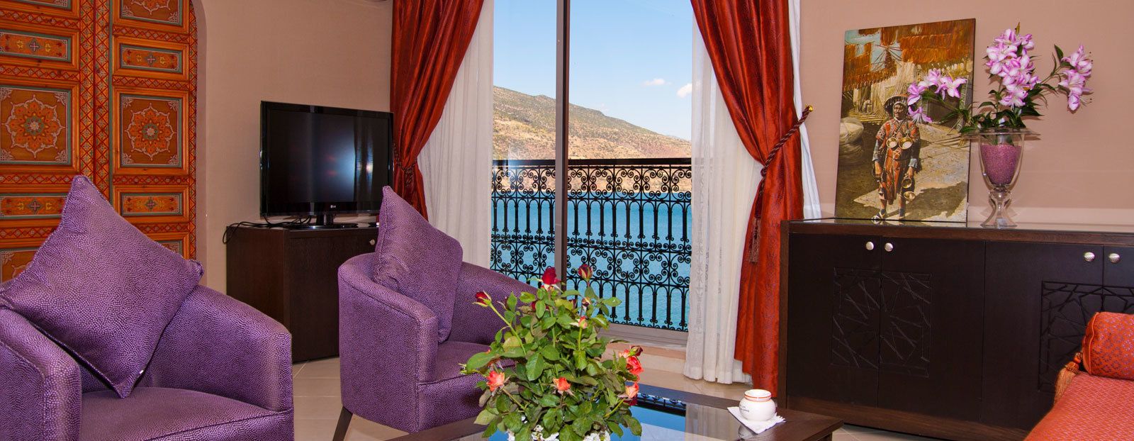 A room with a TV, purple chairs, a table with a vase of flowers, and a view of a blue railing and mountains.