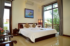 A hotel room with a large bed, a painting on the wall, and a balcony with a view of greenery.