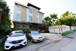 Un hotel con fachada moderna, un estacionamiento con dos coches y un cartel que indica el nombre del hotel.