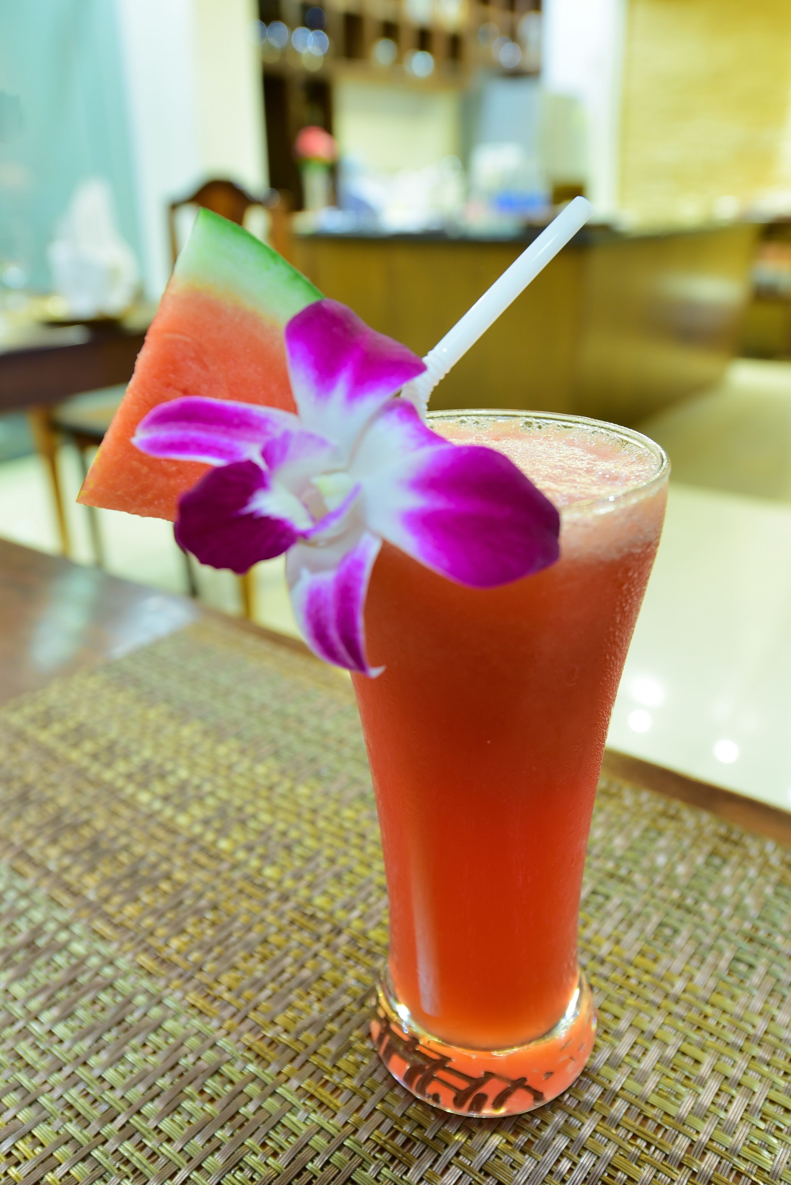 A tall glass with a pink drink, a purple flower, and a slice of watermelon on the rim.