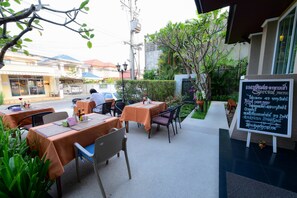 Un café en plein air avec des tables et des chaises, un panneau indiquant les plats proposés, et une vue sur la rue et les bâtiments en arrière-plan.
