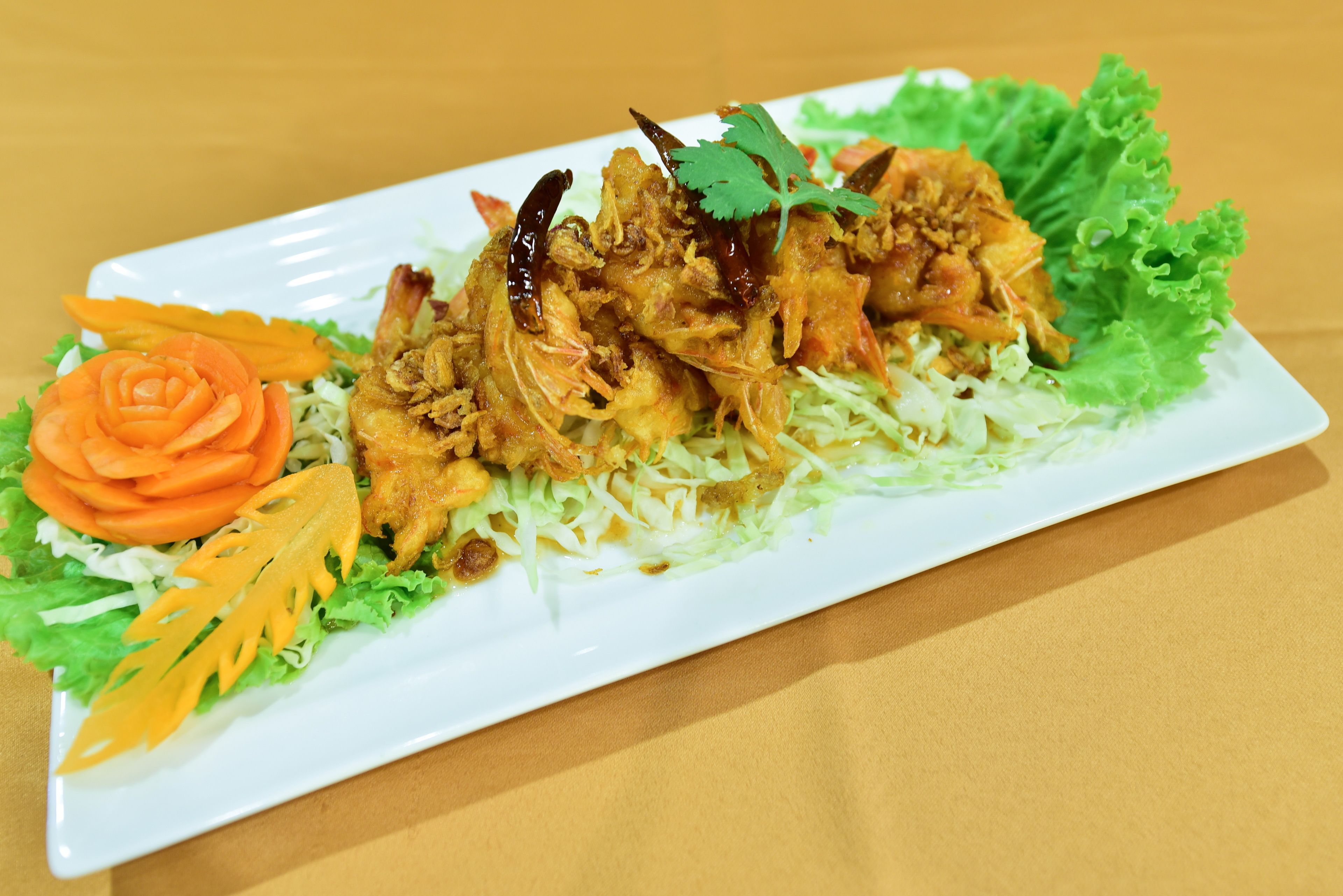 A rectangular plate with a dish of fried chicken garnished with cilantro, red chili peppers, and shredded vegetables on a bed of lettuce.