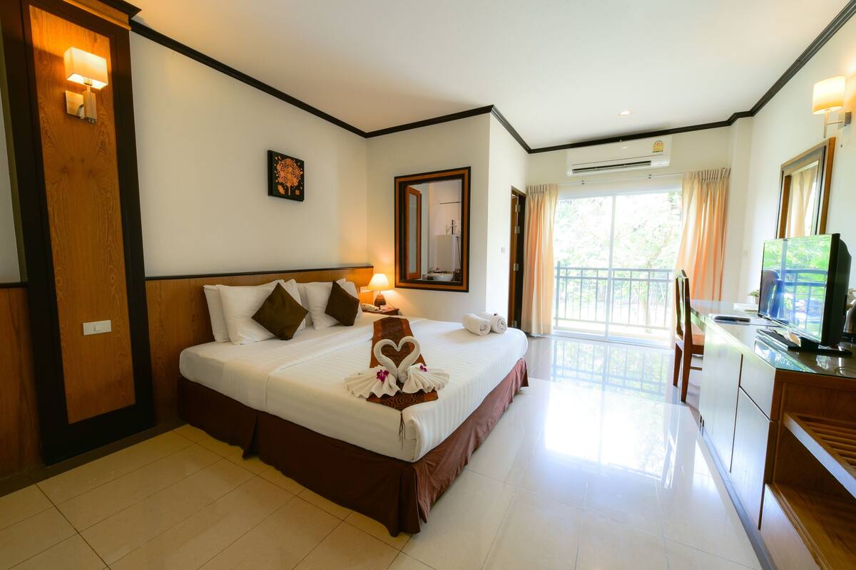 A hotel room with a large bed, a desk with a TV, and a balcony with curtains.