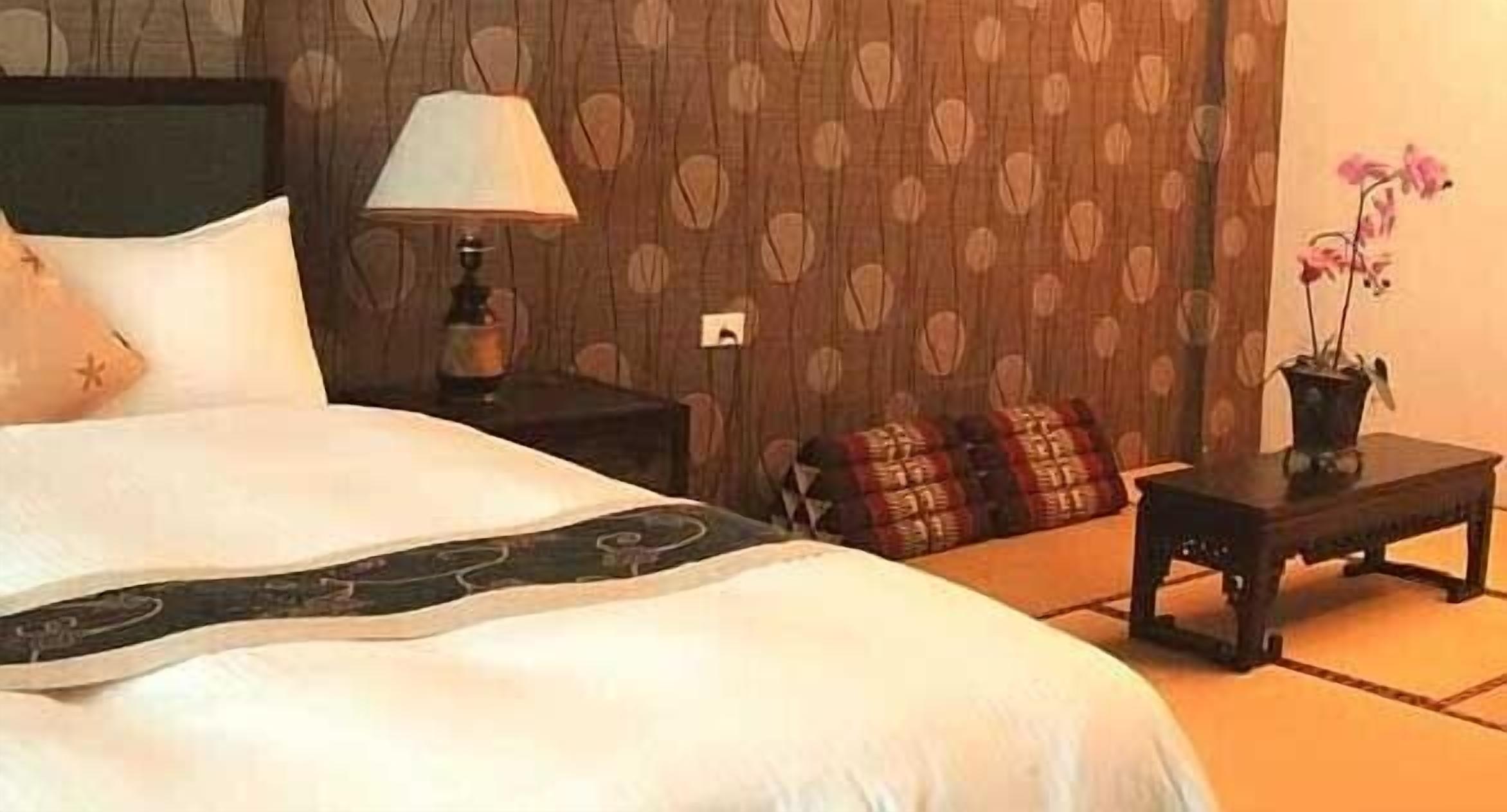 A neatly made bed with a white duvet, a bedside lamp, a small table with a potted plant, and a patterned wall.