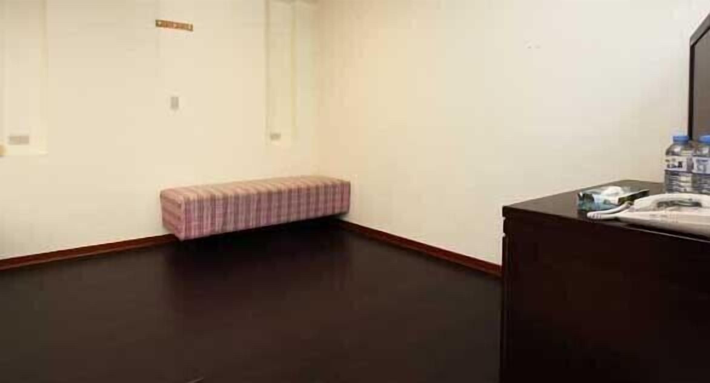 Une chambre d’hôtel avec un plancher en bois foncé, un petit banc rose et un bureau en bois foncé sur lequel se trouvent un téléphone et une bouteille d’eau.