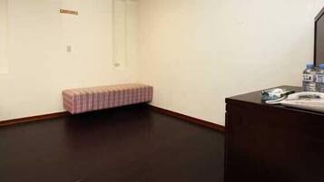 Une chambre d’hôtel avec un plancher en bois foncé, un petit banc rose et un bureau en bois foncé sur lequel se trouvent un téléphone et une bouteille d’eau.
