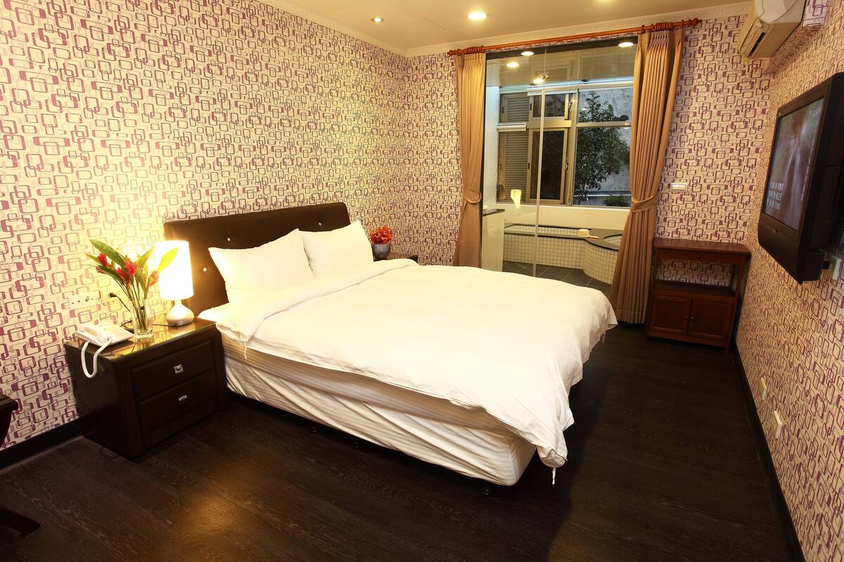 Una cama bien tendida con edredón blanco, una mesita de noche con lámpara y teléfono, y un televisor montado en la pared.