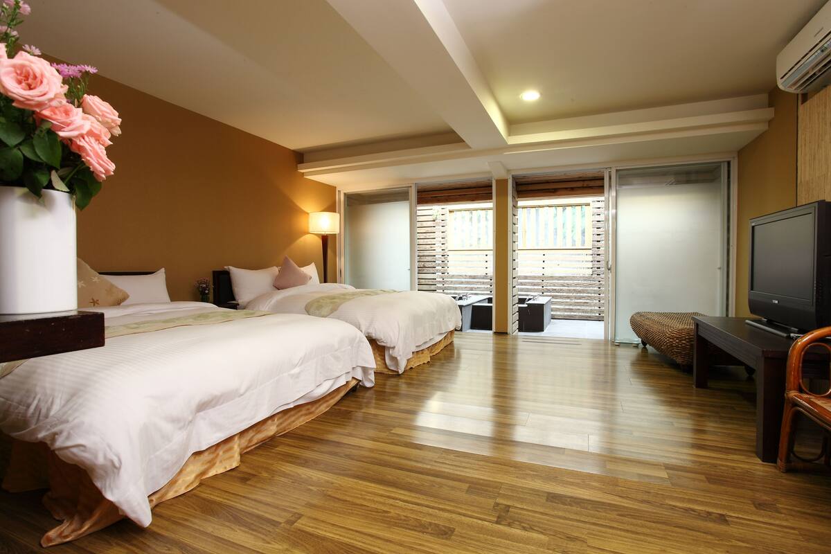 Habitación de hotel con dos camas, televisión, piso de madera y puerta corredera que da a un balcón.