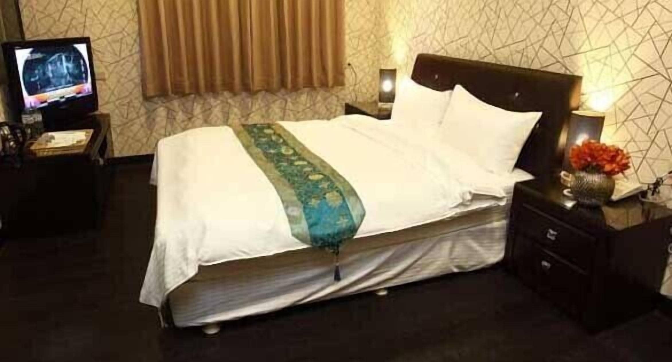 Una cama bien hecha con colcha blanca y verde, cabecera oscura y mesita de noche con un florero.