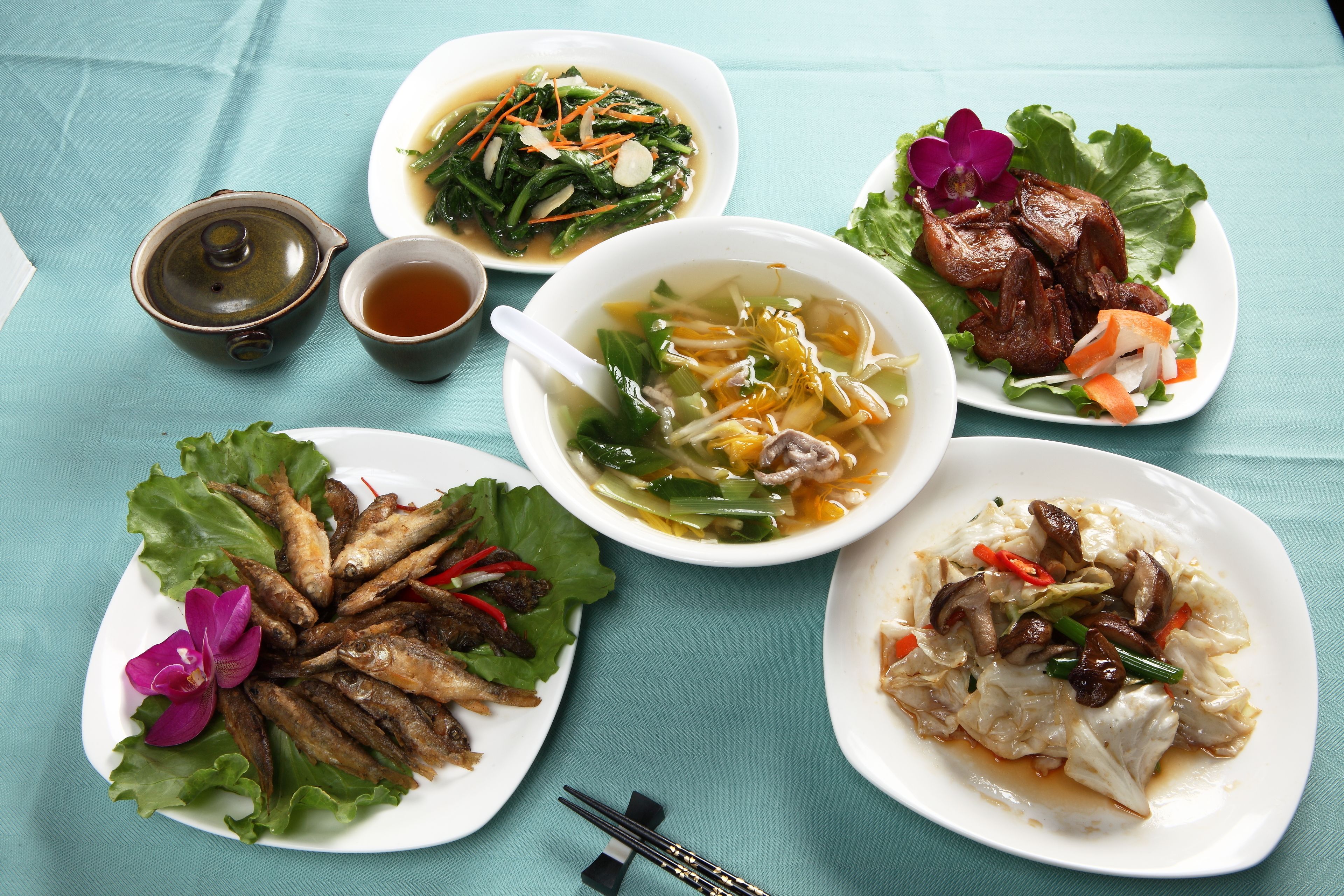 A table with various dishes including soup, grilled fish, and meat, accompanied by a teapot and cups.