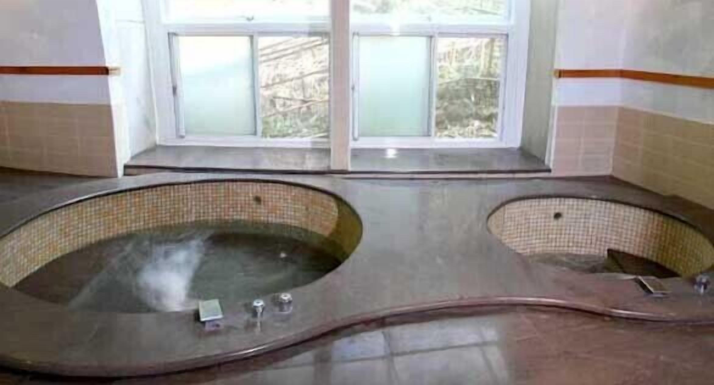 Una habitación con dos tinas de hidromasaje grandes y ovaladas, con interiores revestidos de azulejos, situadas bajo un ventanal con vidrios esmerilados.