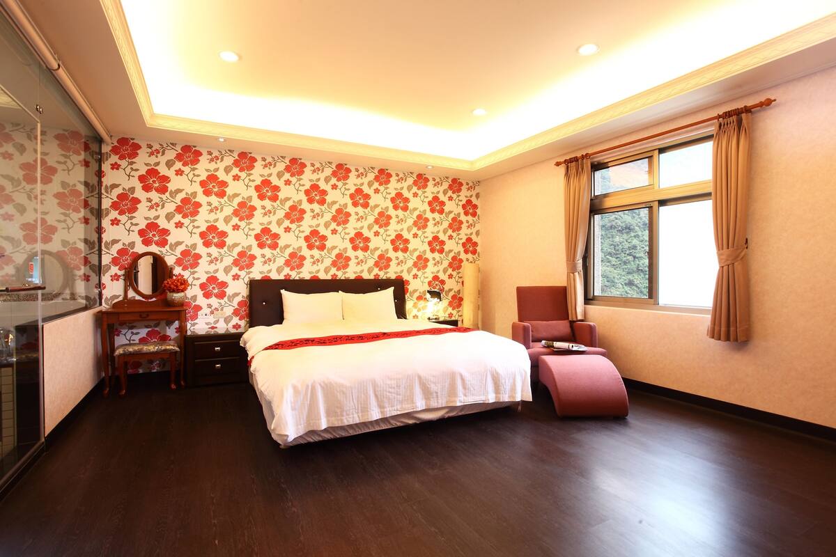 Un dormitorio con una cama grande, una mesita de noche, una silla y una ventana con cortinas.