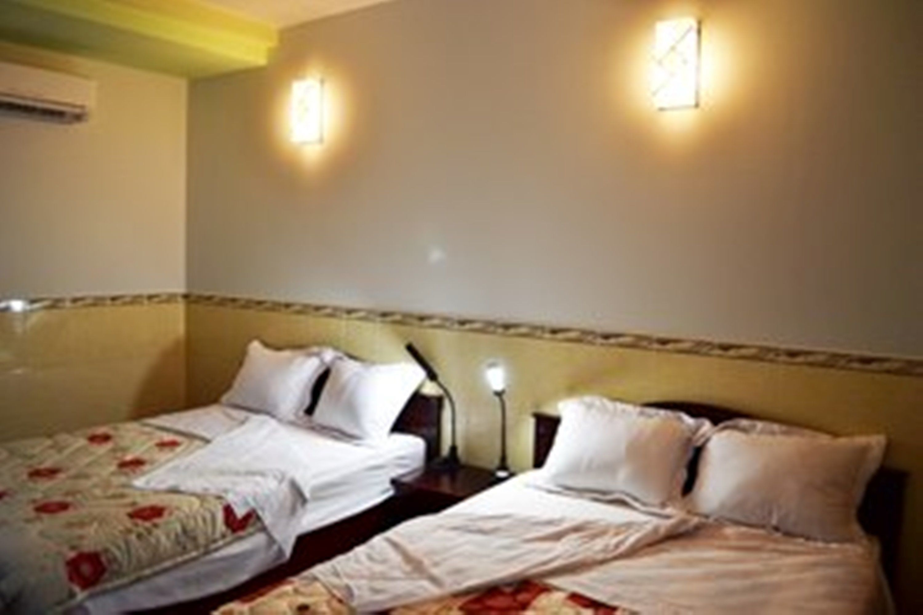 Habitación de hotel con dos camas, cada una con ropa blanca y colchas de estampado floral. Hay lámparas de noche y un aire acondicionado instalado en la pared.