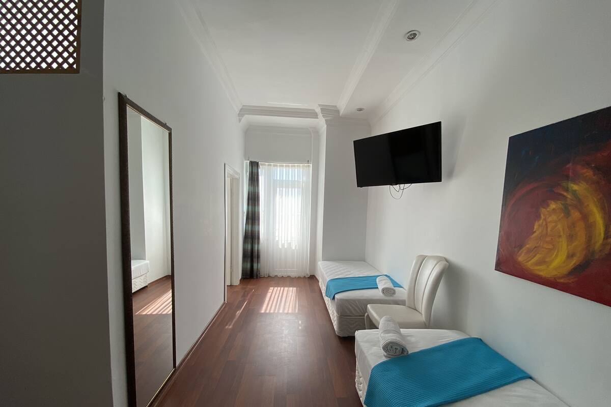 A hotel room with a wooden floor, a bed with a blue blanket, a white sofa, a TV mounted on the wall, and a painting.