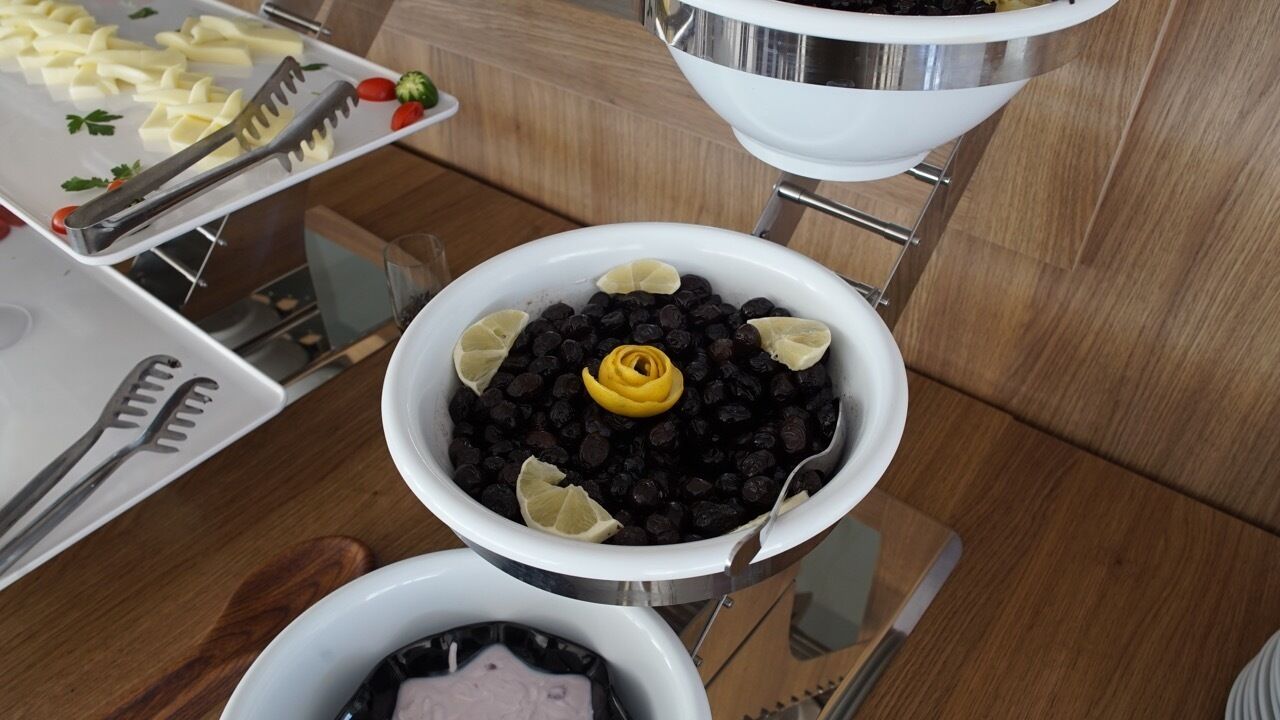 A buffet setup with various dishes, including a bowl of black olives garnished with lemon slices and a decorative yellow flower.