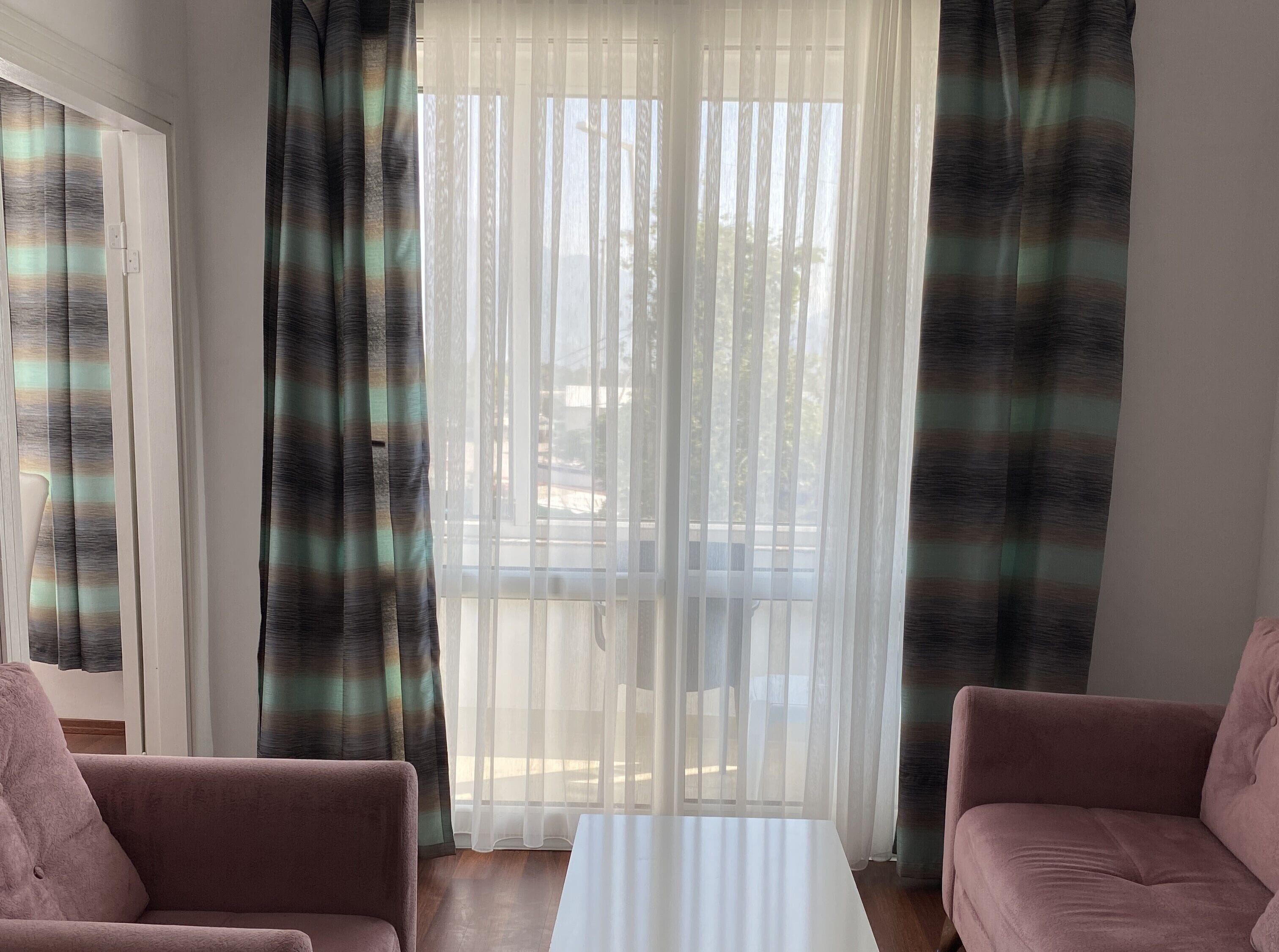 Ένα δωμάτιο με έναν καναπέ, ένα τραπέζι και θέα από ένα παράθυρο με λεπτά, διάφανα κουρτίνια.