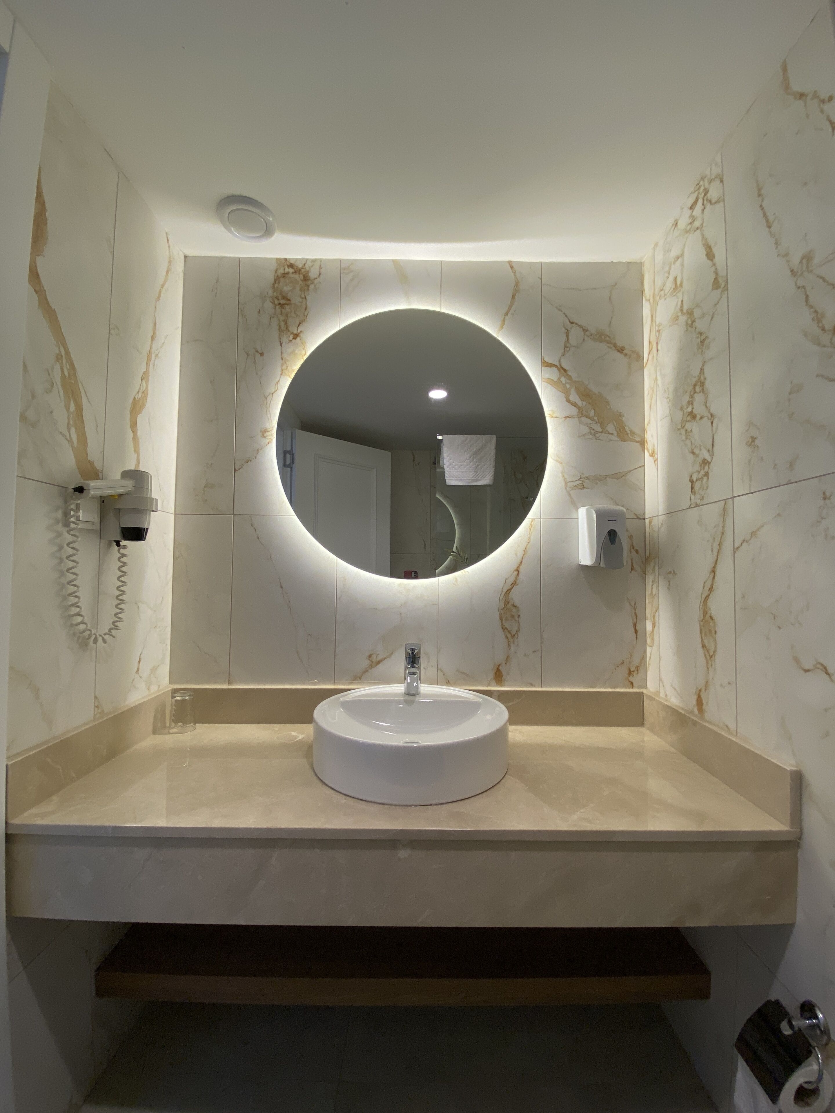 A bathroom with a round mirror, a sink, and a towel dispenser.