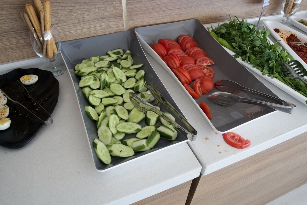 A kitchen counter with trays of sliced cucumbers, tomatoes, and herbs.