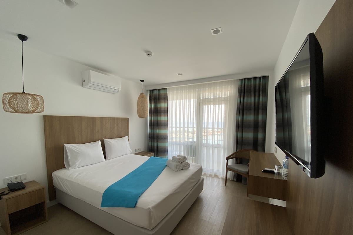 A modern hotel room with a large bed, a flat-screen TV, a wooden nightstand, and a view of the beach through the window.