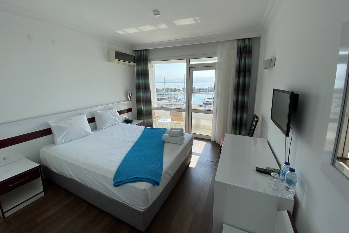 A hotel room with a bed, a desk with a TV, a chair, and a view of the marina through the window.