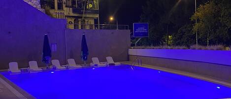 An outdoor swimming pool at night with blue lighting, a stone wall, and a building with a sign for an alcohol shop and tobacco shop.