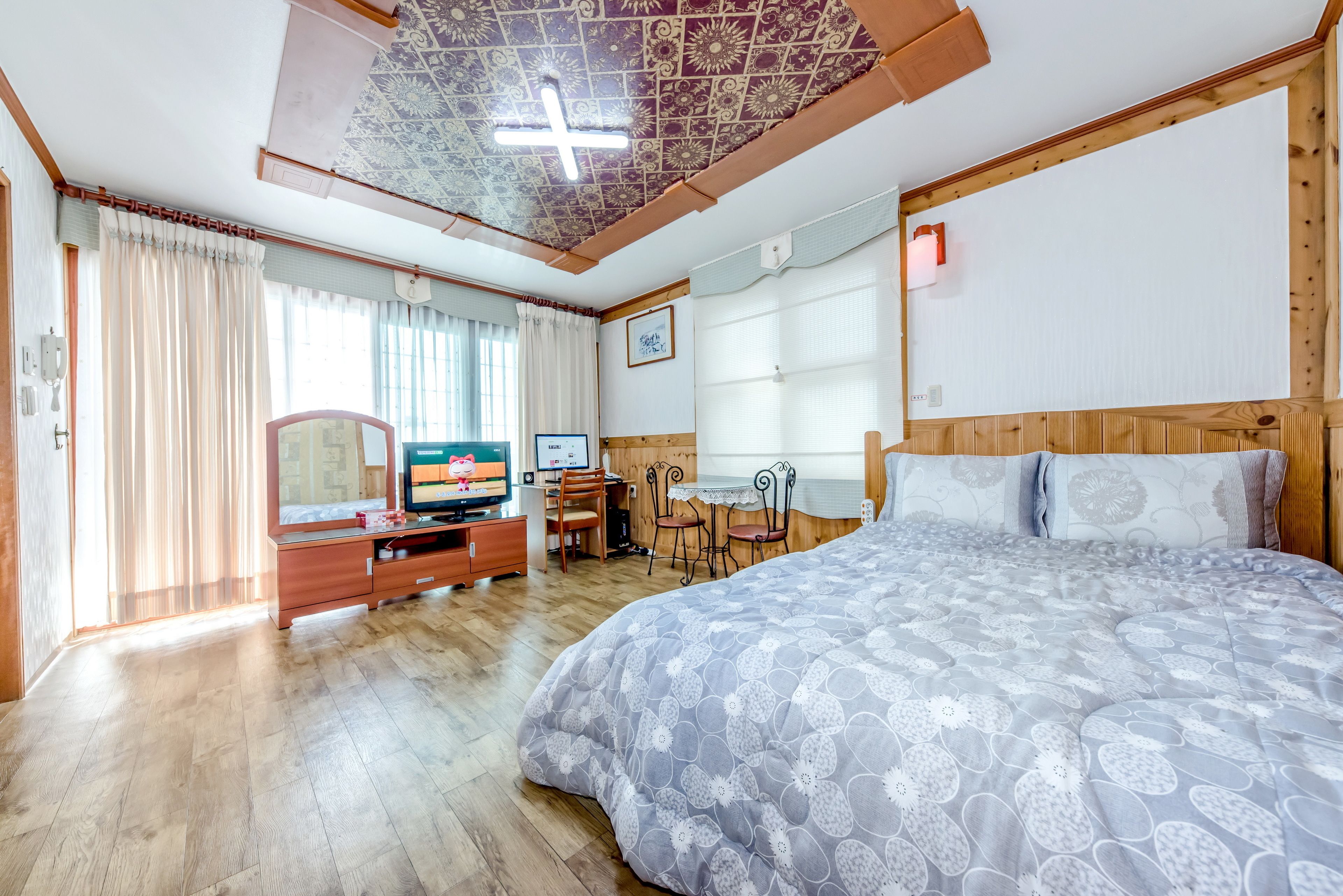 A bedroom with a large bed, wooden headboard, a desk with a chair, a television, and a window with curtains.