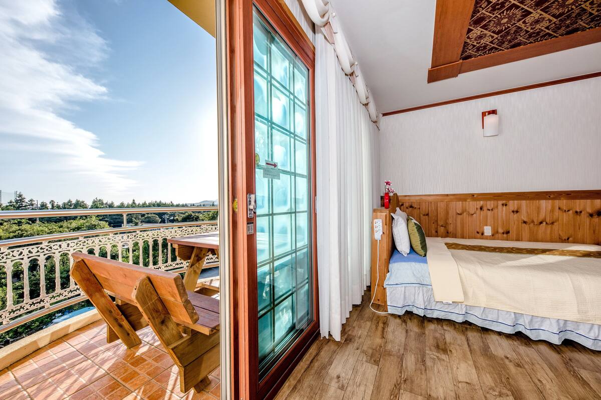 A balcony with a wooden table and chairs, a bed with a wooden headboard, and a view of greenery.