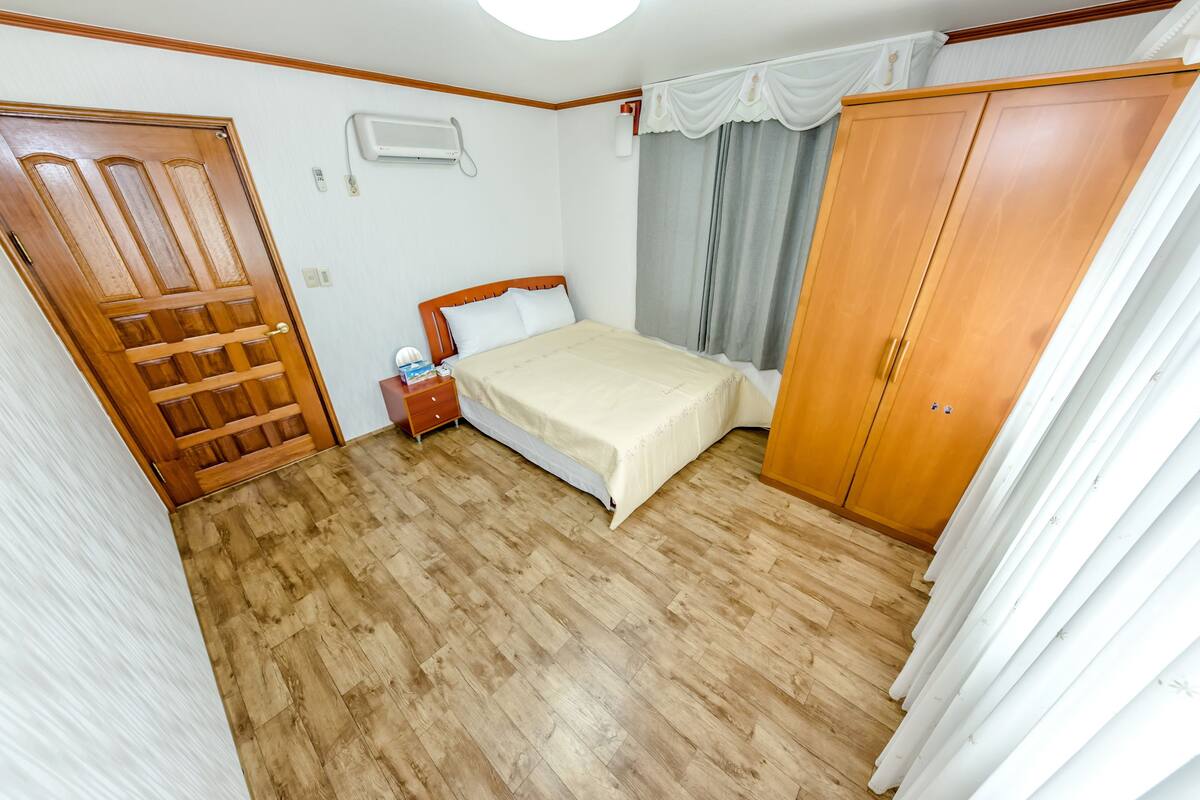 A bedroom with a wooden floor, a bed with a white bedspread, a wooden wardrobe, and a window with curtains.