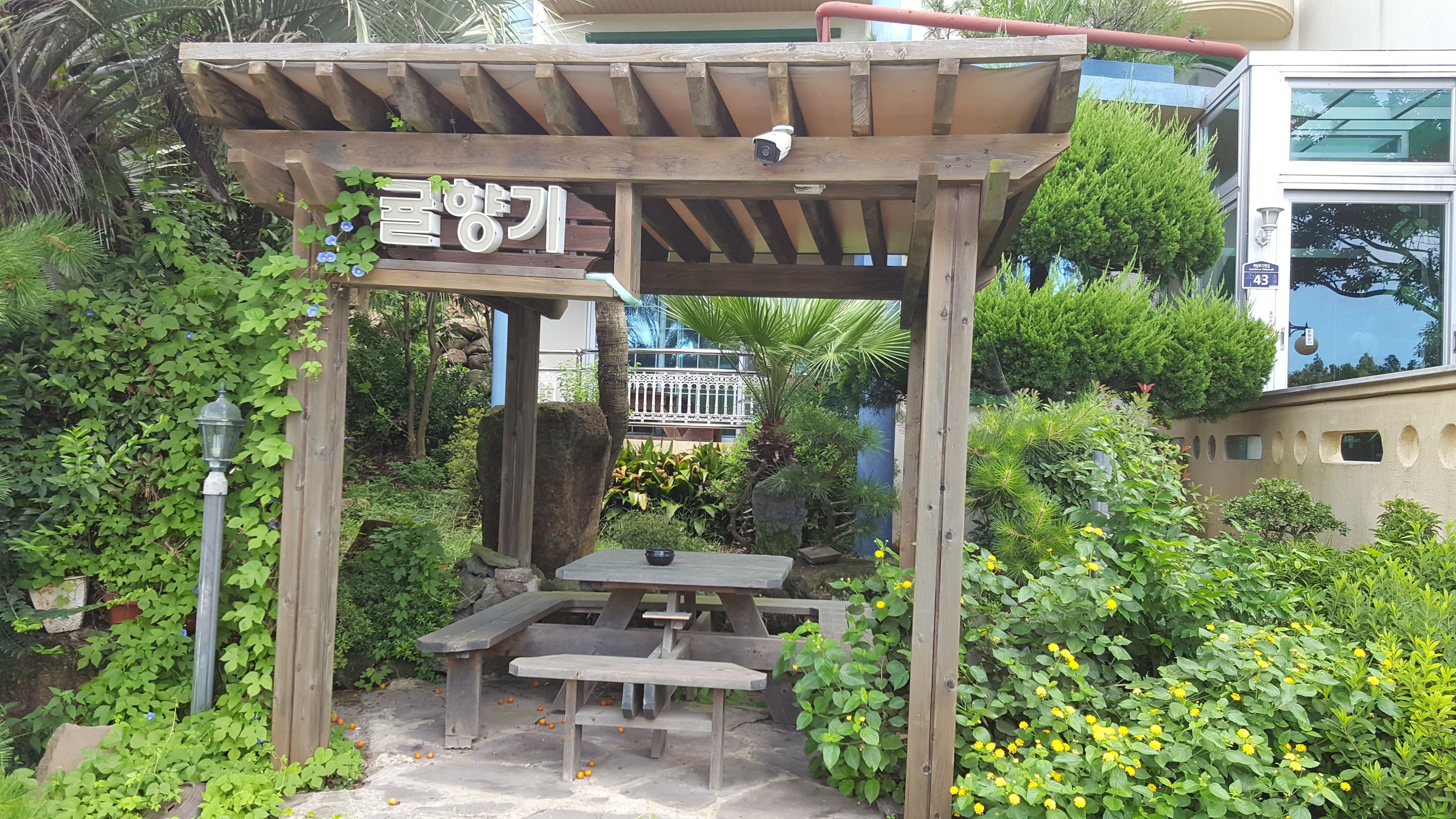 A wooden pergola with a sign, a table, and benches in a garden area with various plants and a building in the background.