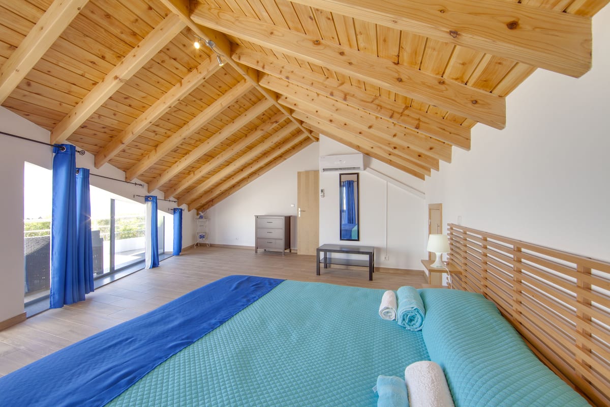 A spacious bedroom with a high wooden ceiling, a large bed with blue bedding, a bedside table, and a view through a window with blue curtains.