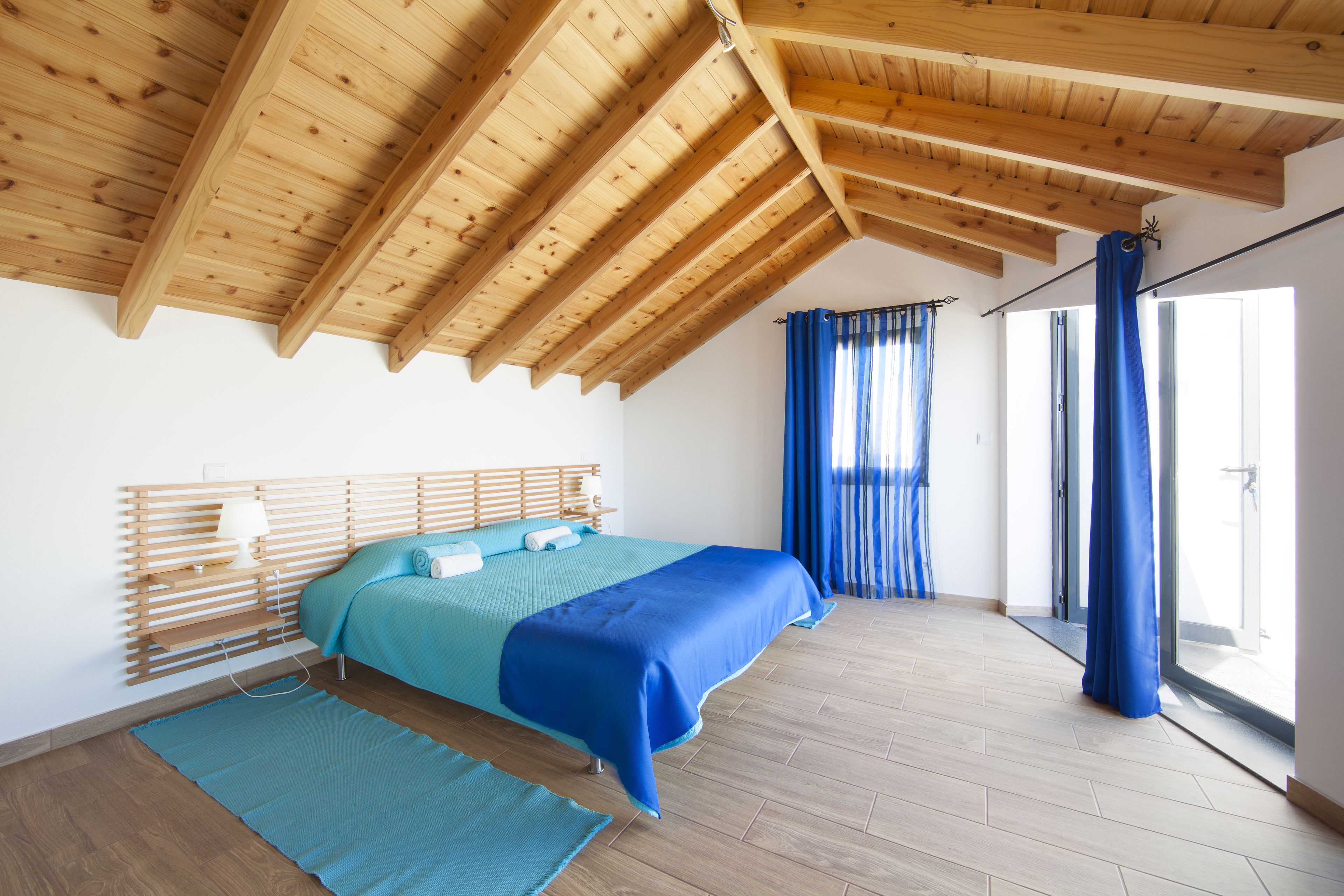 A bedroom with a wooden ceiling, a bed with blue bedding, a wooden headboard, and a large window with blue curtains.