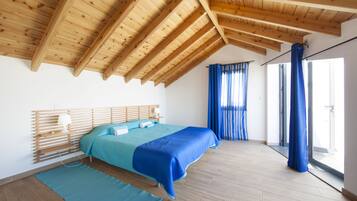 A bedroom with a wooden ceiling, a bed with blue bedding, a wooden headboard, and a large window with blue curtains.