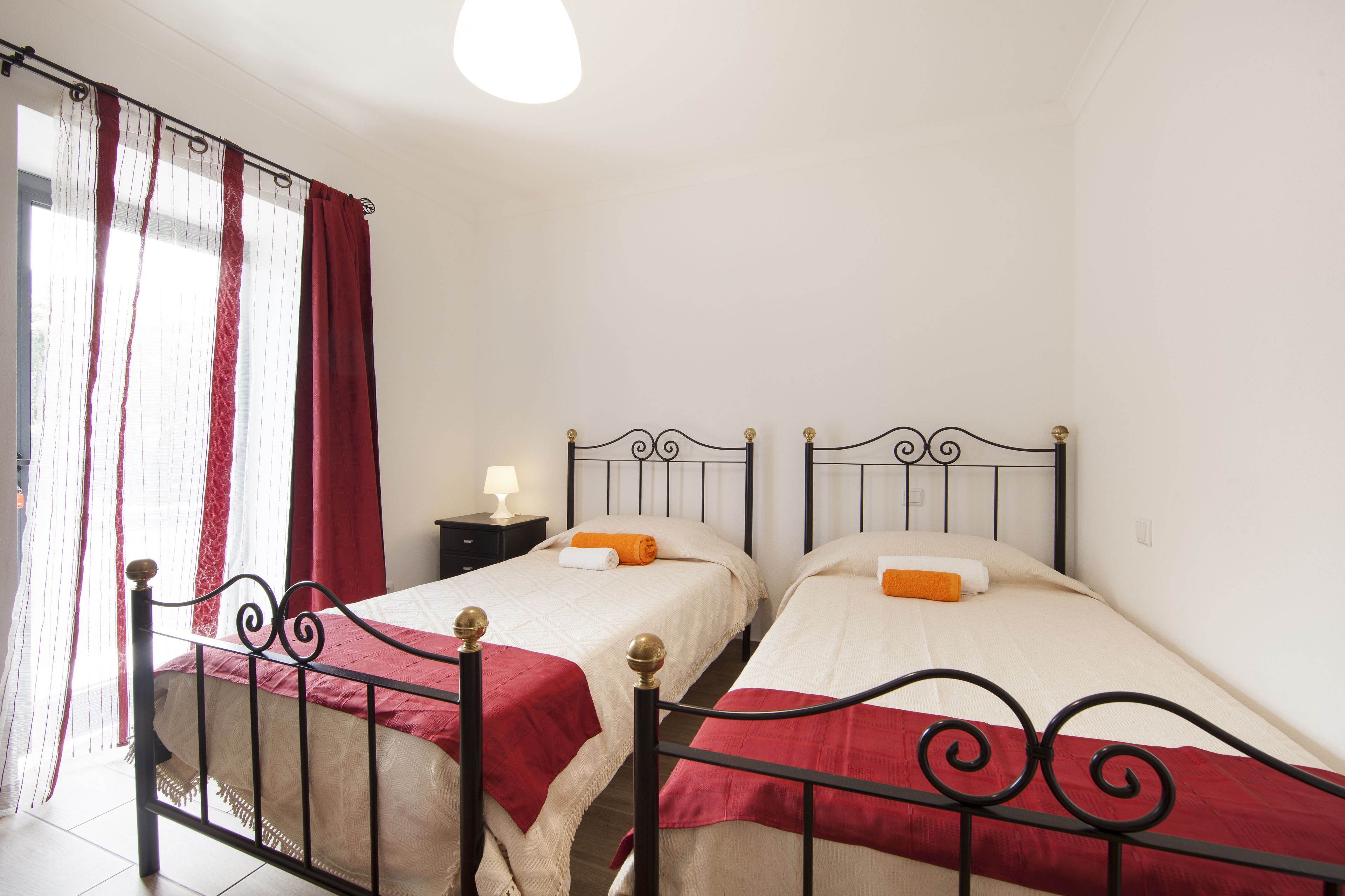 Una habitación con dos camas, cada una con cabecera roja y ropa de cama blanca. Hay una mesita de noche con una lámpara entre las camas.