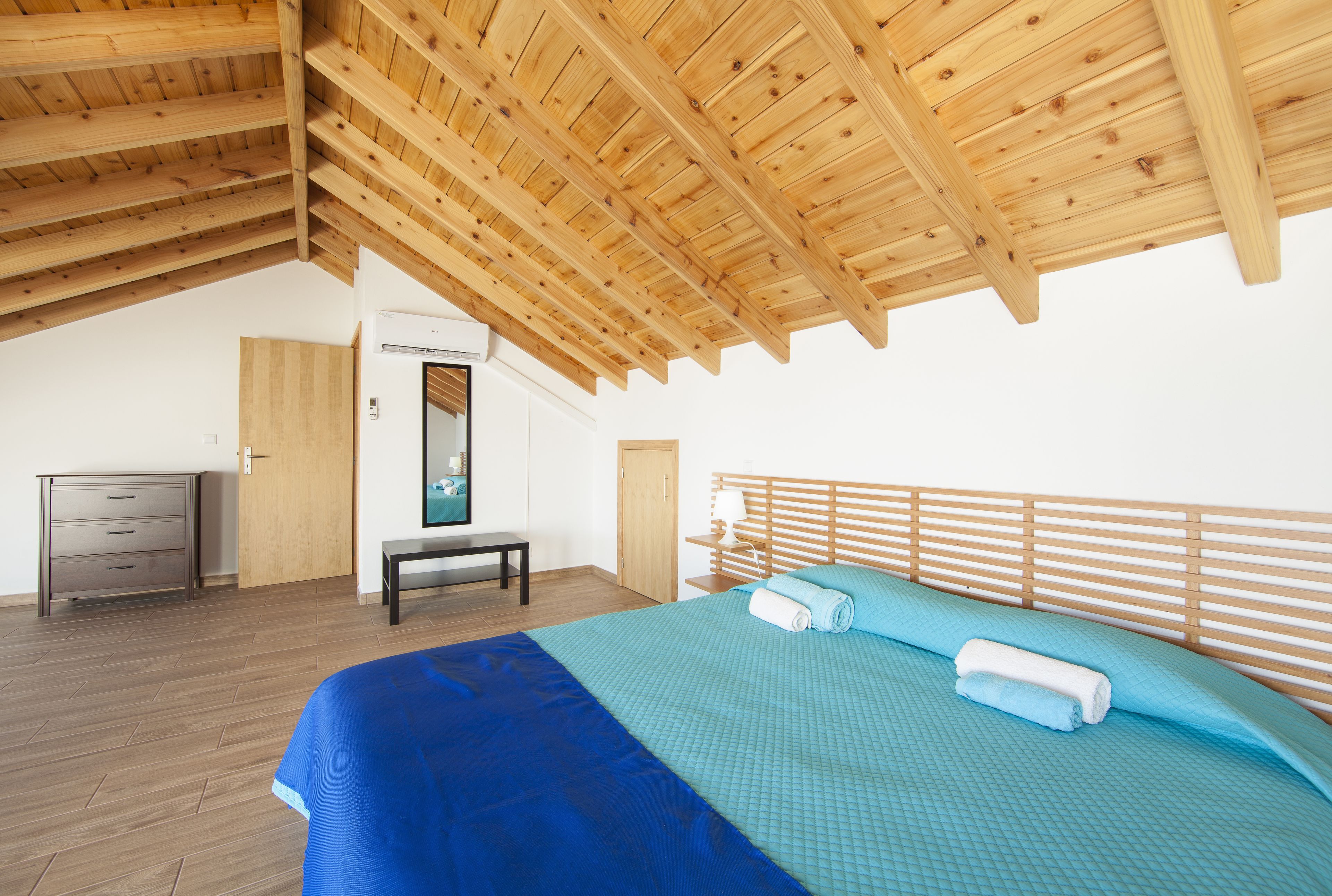 Un dormitorio con techo de madera, cama con ropa de cama azul, cabecero de madera, mesita de noche y armario.