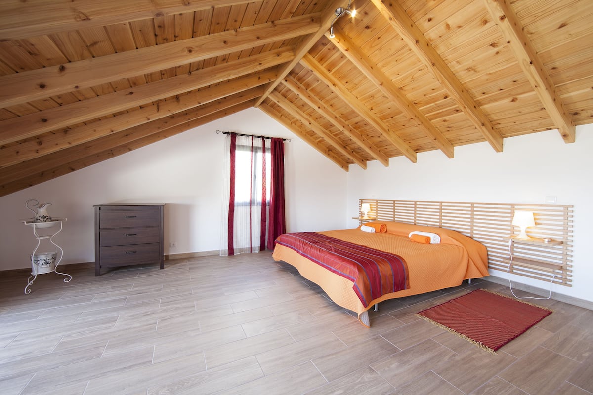 A bedroom with a sloped ceiling, a bed with an orange bedspread, a bedside table with a lamp, and a small dresser.